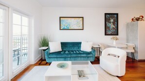 A modern living room with a teal sofa, white armchairs, and a glass coffee table. There are two framed artworks on the wall, a potted plant, and a view of the city through large windows.