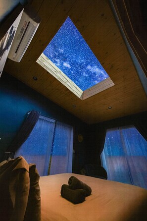 A room with a skylight displaying a starry night sky, a bed with a blanket, and a wooden ceiling.
