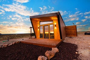 A modern wooden cabin with a large glass door and windows, situated on a gravel ground with a clear sky in the background.