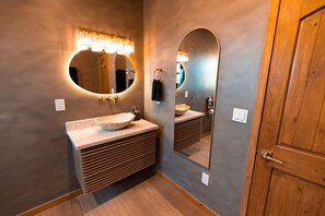 A bathroom with a wooden vanity, a round mirror with a lighted frame, and a wooden door.