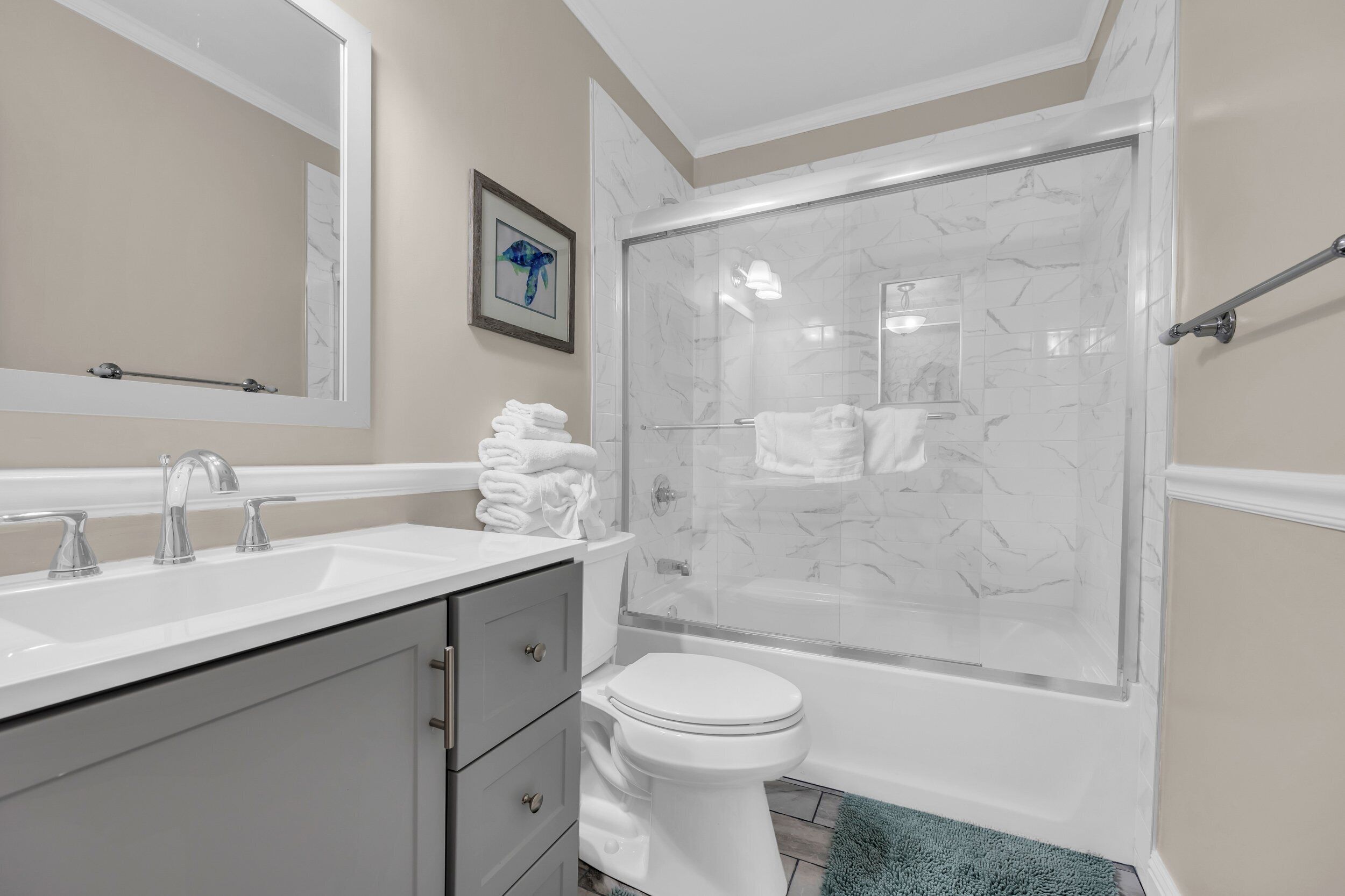 A modern bathroom with a white sink, vanity, and toilet. It includes a glass shower enclosure and a framed artwork on the wall.