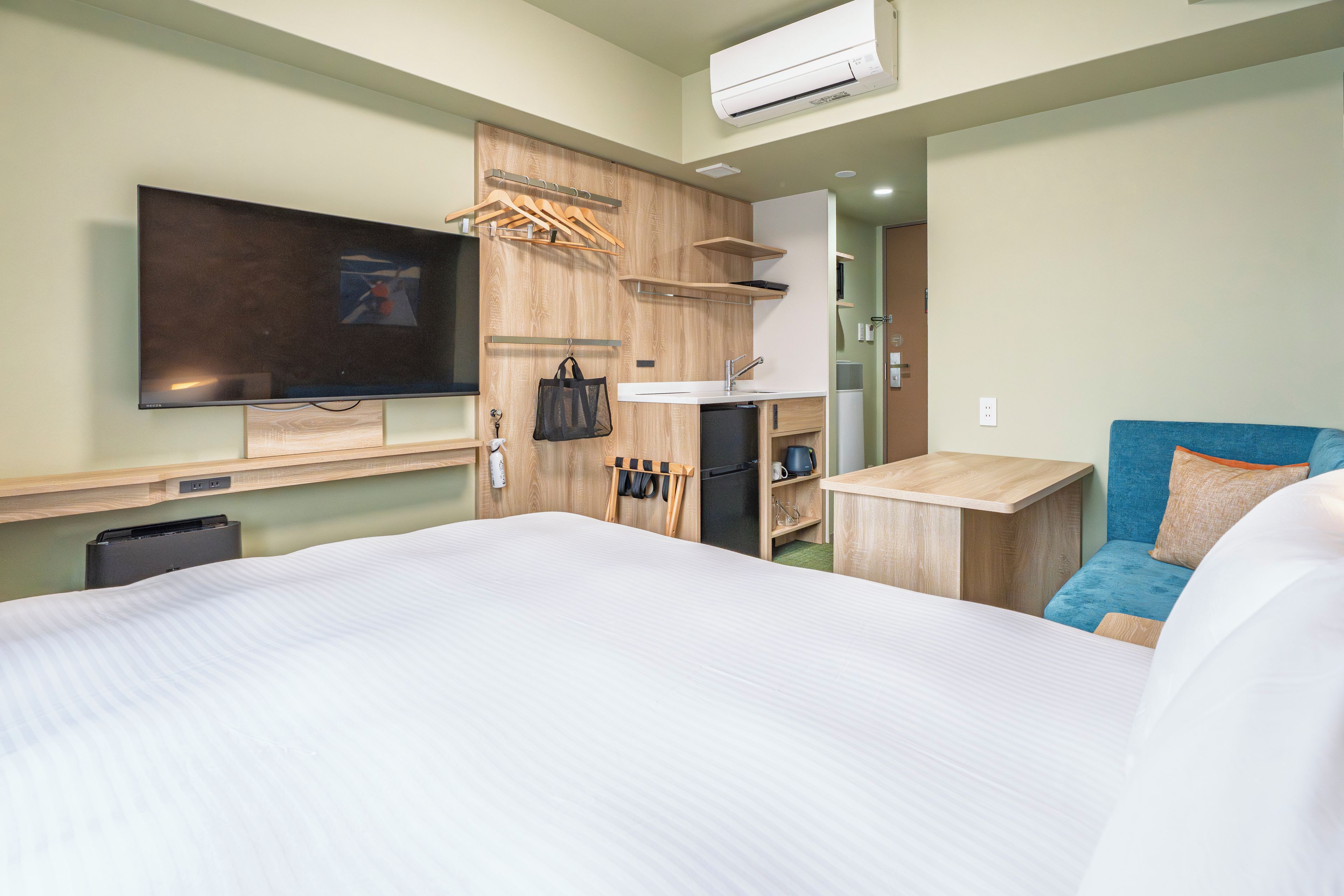 Una habitación de hotel con una cama, un televisor montado en la pared, una kitchenette pequeña y un sofá azul.