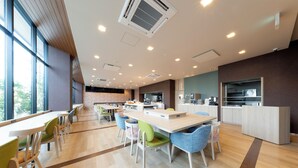 A modern dining area with multiple tables and chairs, a kitchen area, and large windows providing natural light.