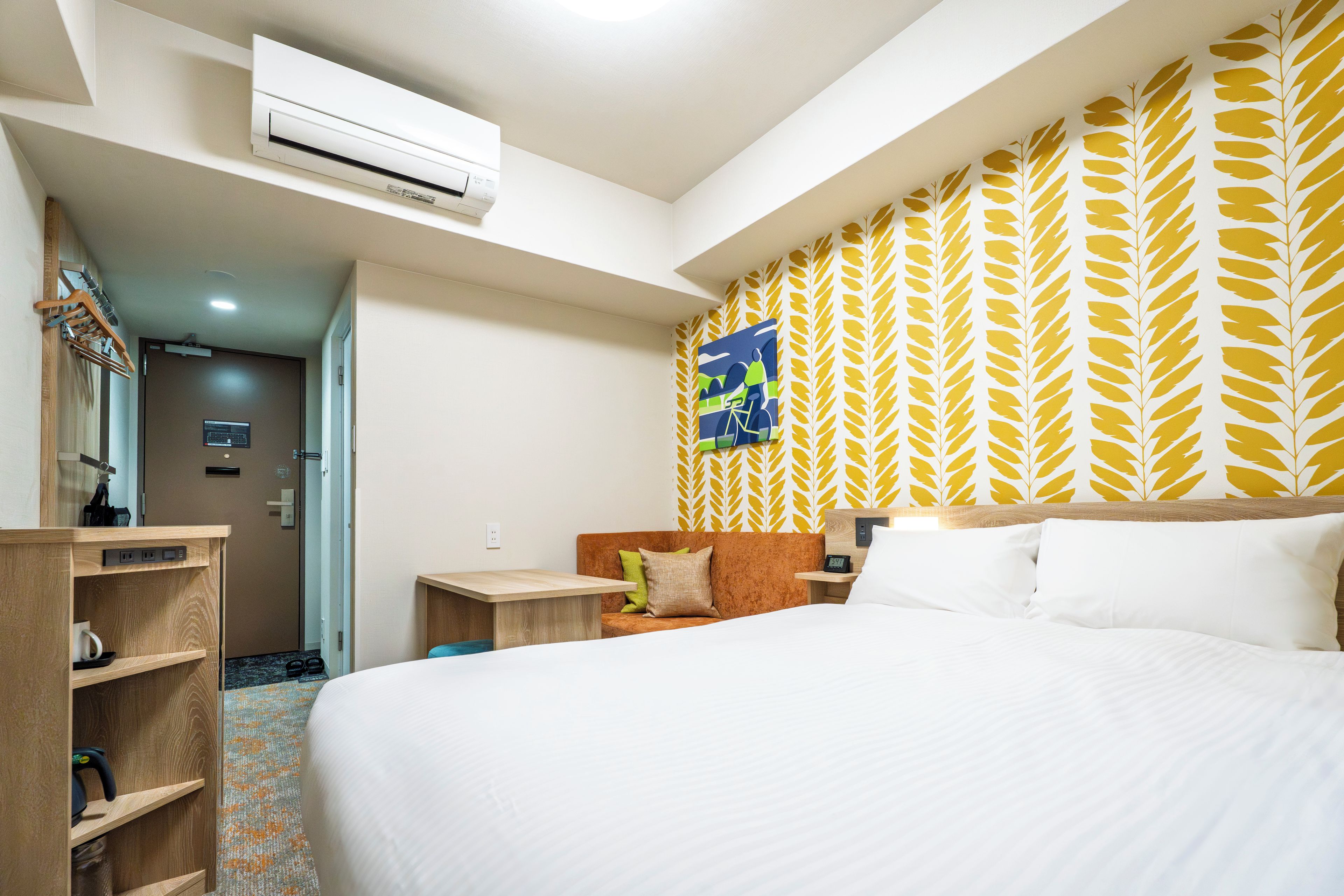 A hotel room with a bed, a desk, a chair, a wardrobe, an air conditioning unit, and a patterned wall.