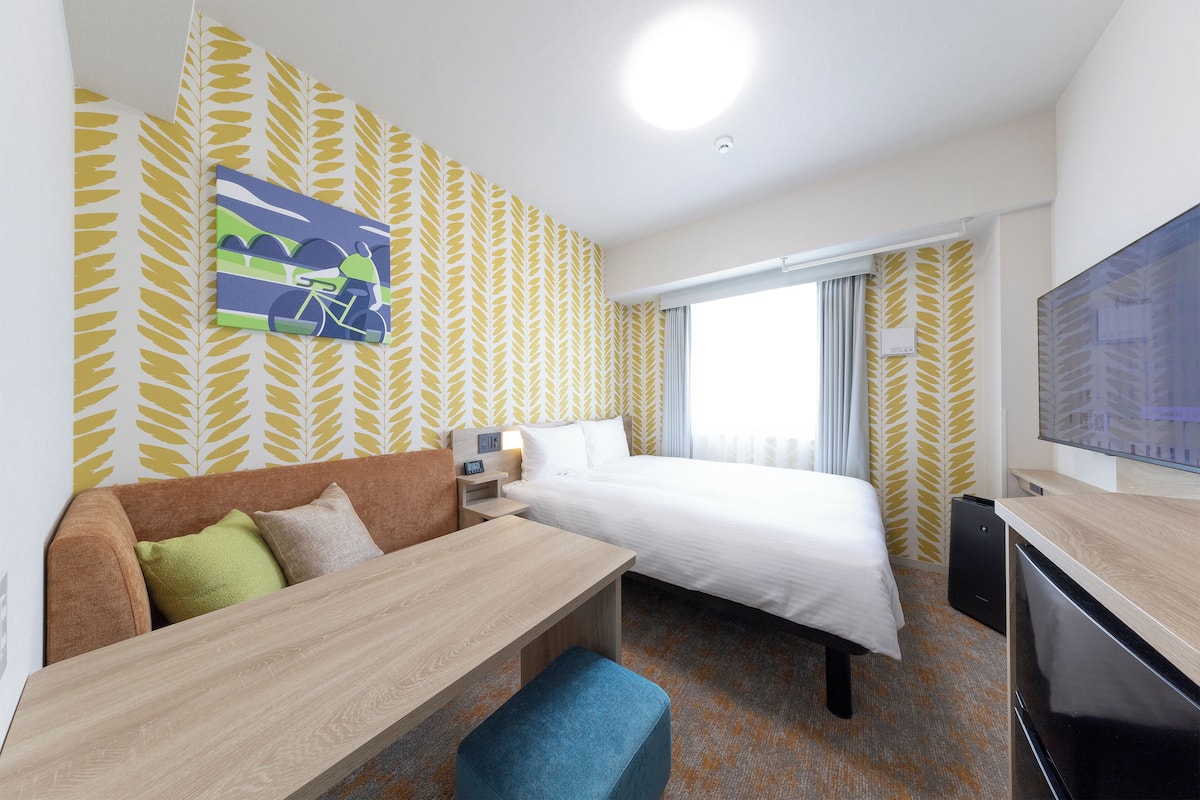 Ein Hotelzimmer mit Bett, einer Sitzecke, einem Schreibtisch, einem Fernseher und einem fensterbepflanzten Fenster.