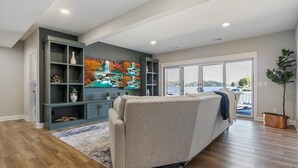A modern living room with a sofa, a bookshelf, and a large window offering a view of the outdoors. - Accessible Lakeside Luxury at the 45mm w/ Private Dock, Sunset Views & More (Climax Springs)