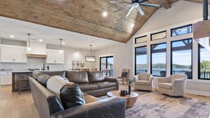A spacious living area with a leather sofa, a coffee table, and two armchairs. A large window offers a view of the outdoors. - Accessible Lakeside Luxury at the 45mm w/ Private Dock, Sunset Views & More (Climax Springs)