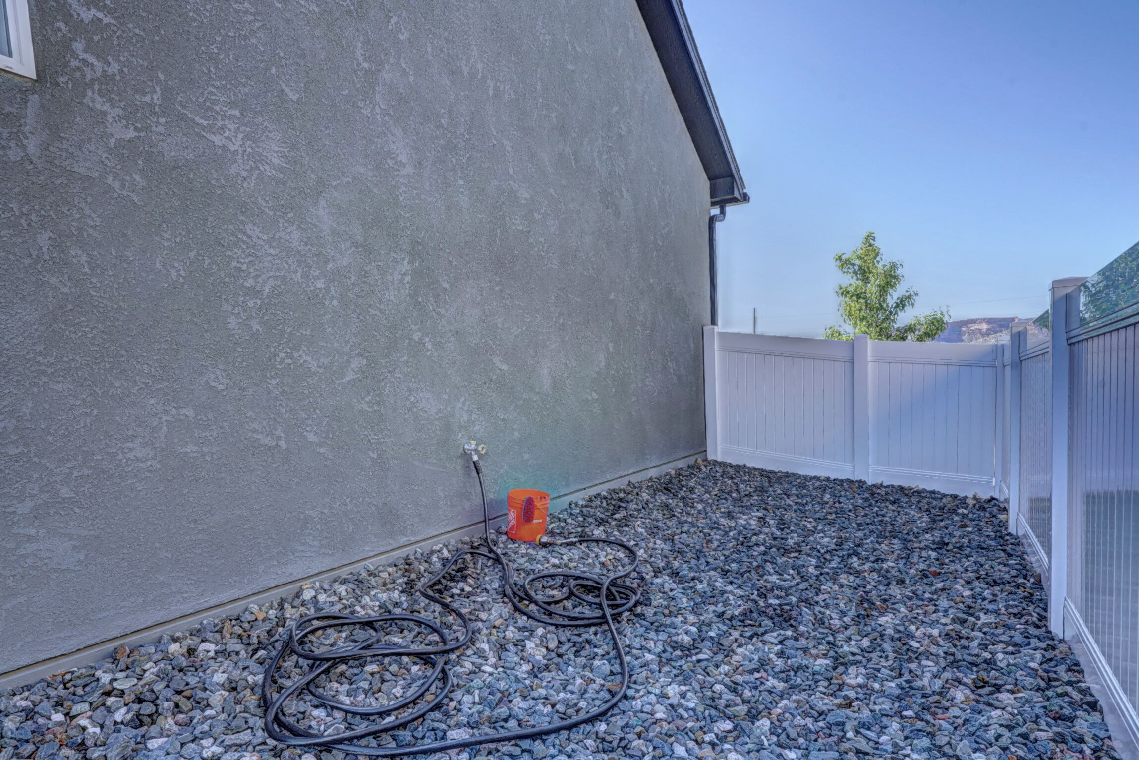 Een achtertuin met een betonnen muur, een wit hekwerk en een tuinslang op een grindpad.