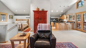 Uma sala de estar aconchegante com uma poltrona de couro, uma mesa de madeira, um armário vermelho e uma área de cozinha visível através de uma porta aberta.