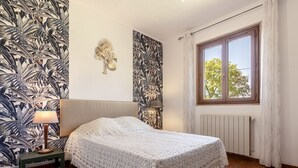 Un dormitorio con una cama, una mesita de noche, una lámpara, un ventanal con cortinas y una pared con estampado de hojas tropicales.