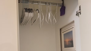 Una habitación con una puerta que da a un armario con ropa colgada y una ventana pequeña con vista al atardecer.