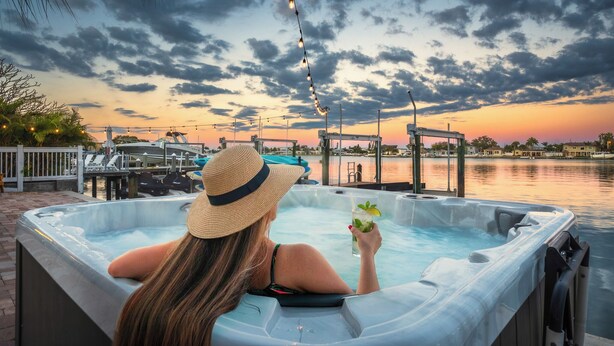 Una persona relajándose en un jacuzzi con una bebida, con vistas a un puerto deportivo con barcos y un atardecer.