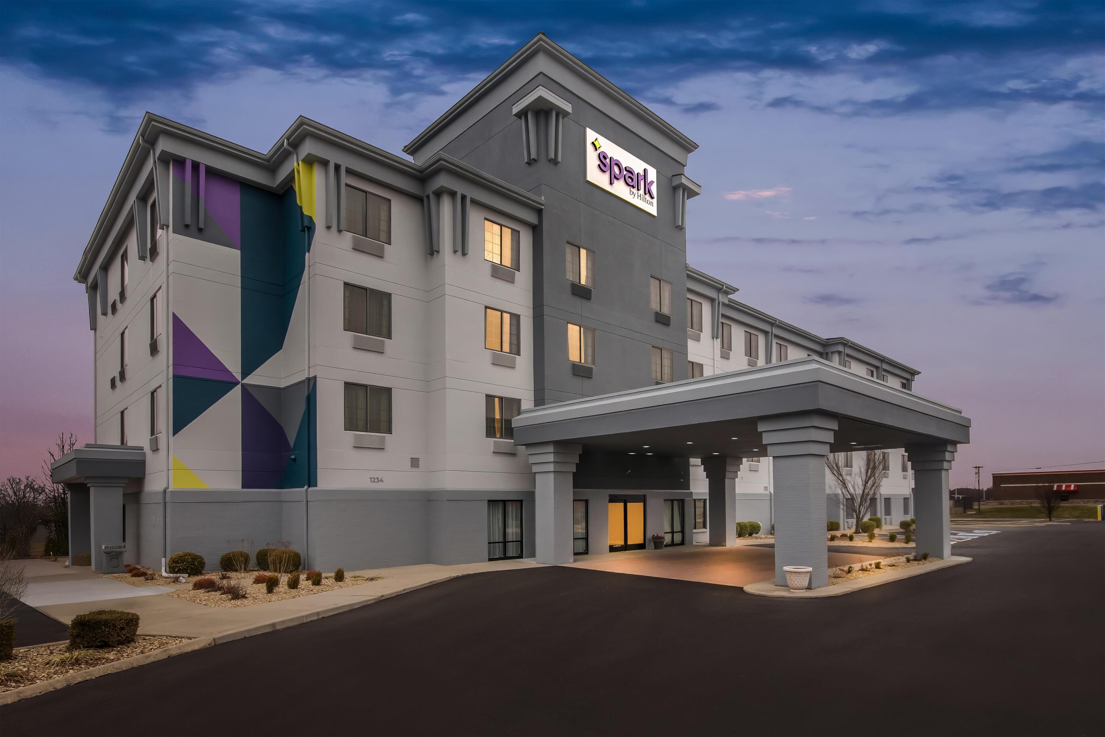 Un hotel de varios pisos con diseño moderno, que cuenta con un llamativo letrero 'spark by Hilton', una entrada cubierta y un camino de acceso arbolado.