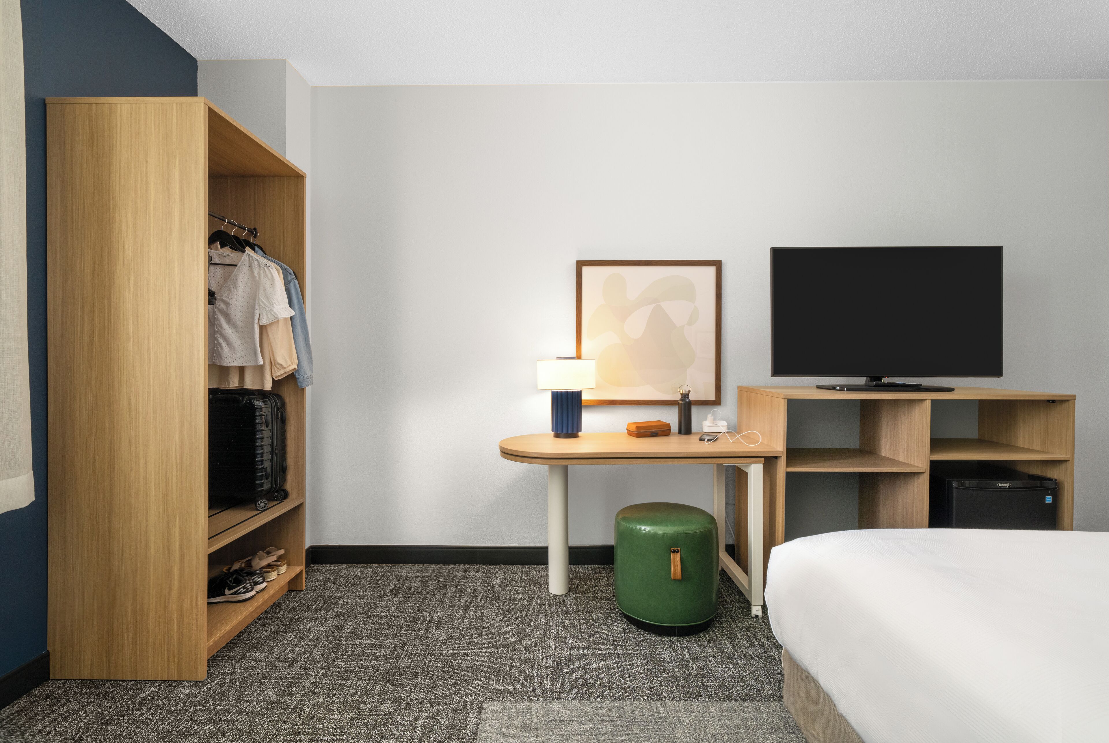Habitación de hotel con cama, armario, escritorio con lámpara, televisor y un cesto de basura.