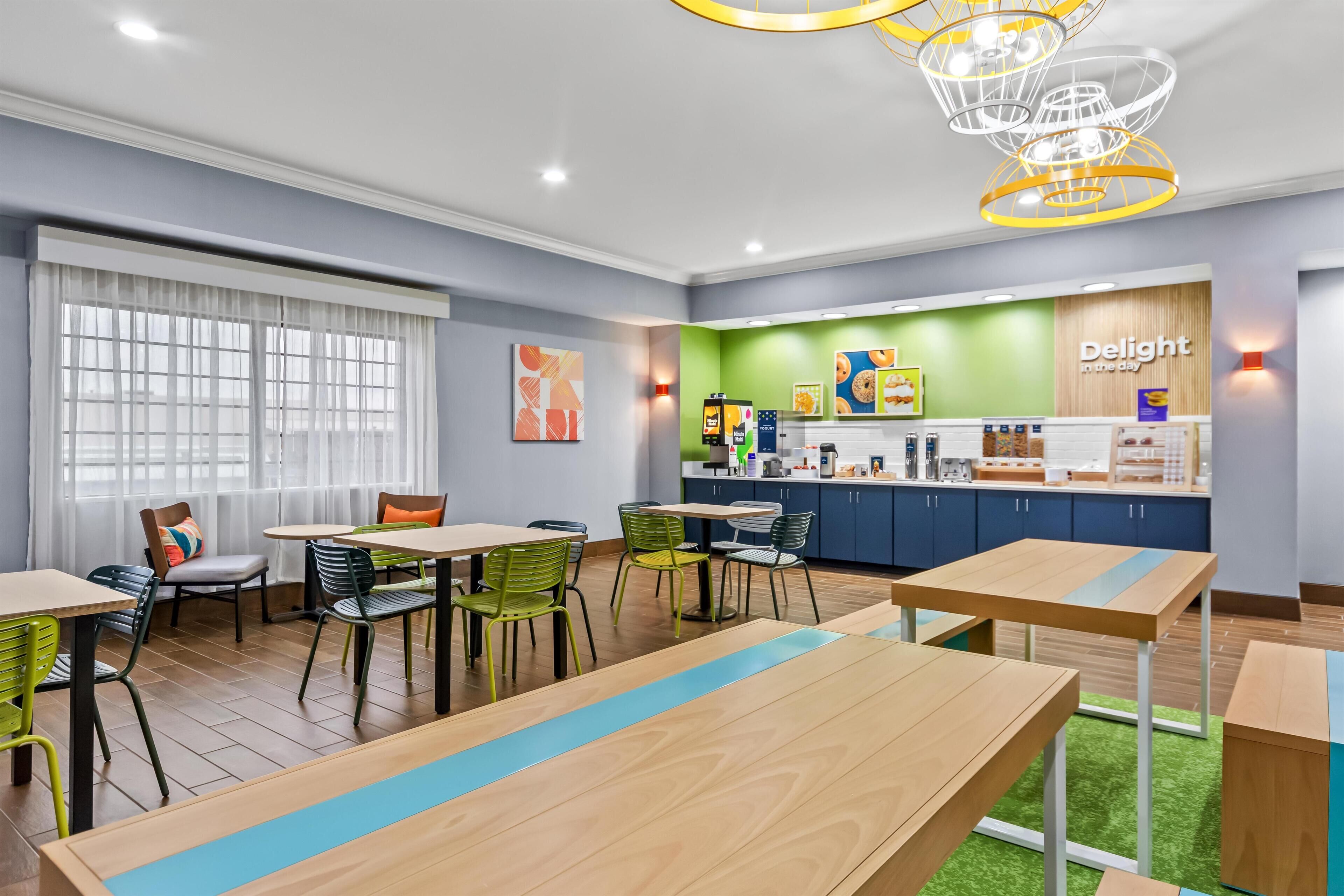 A modern dining area with wooden tables and chairs, a counter with food items, and a green wall with the text "Delight in the day".