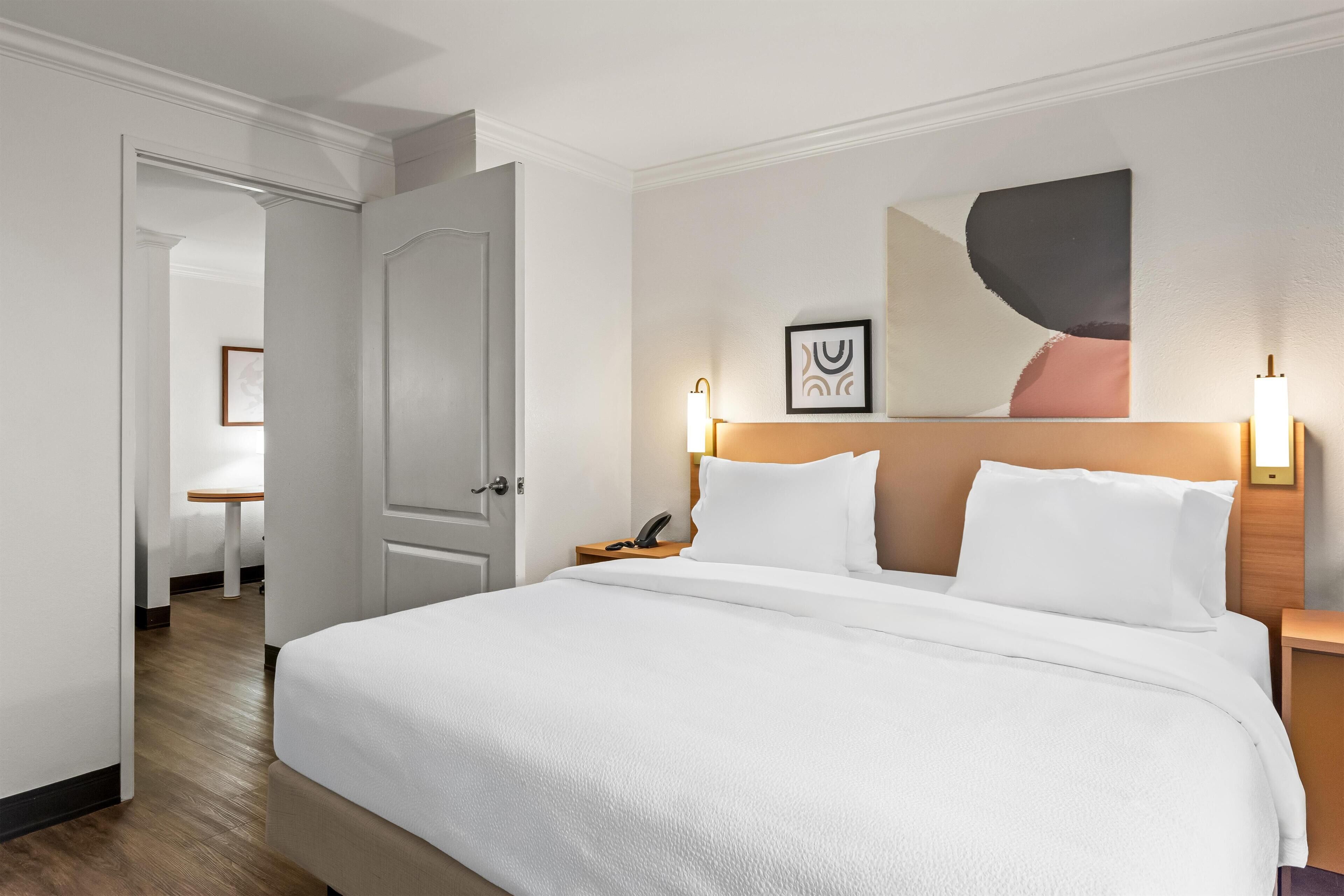 Une chambre d’hôtel avec un grand lit, une table de chevet avec une lampe et une porte donnant sur une autre pièce.
