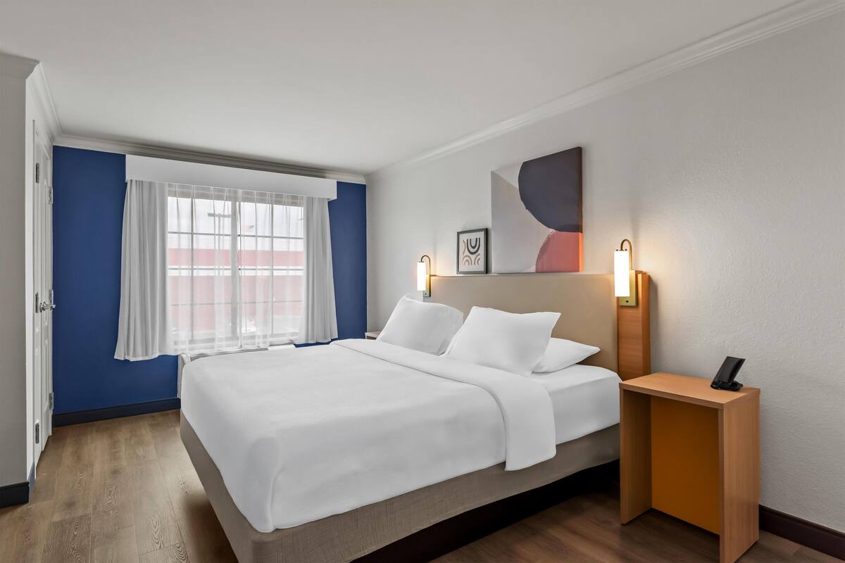 Et hotelværelse med seng, natborde, et vindue med gardiner og et billedværk hængende på væggen.