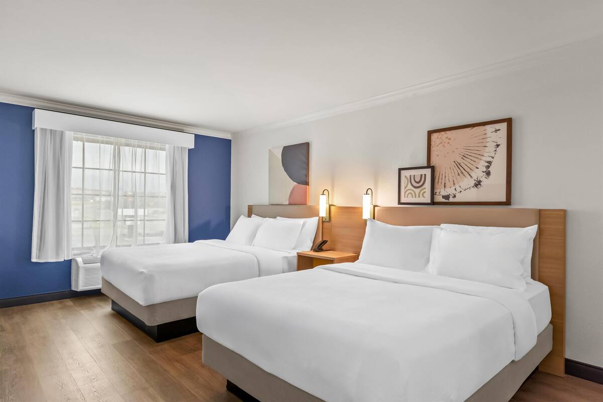 Et hotelværelse med to senge, et stort vindue med hvide gardiner og billeder på væggen.