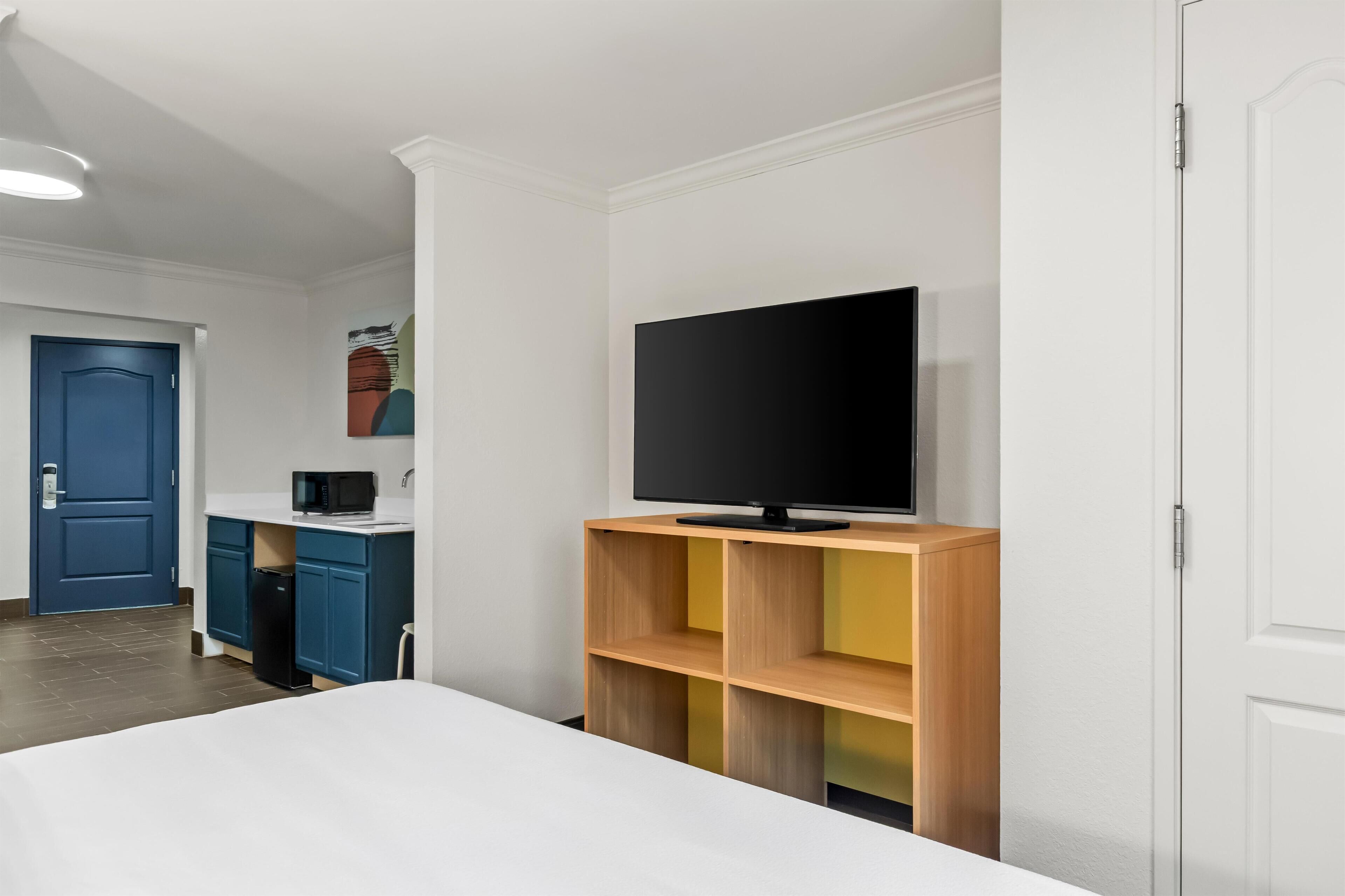 A hotel room with a bed, a TV on a wooden stand, a kitchenette with a microwave, and a blue door.