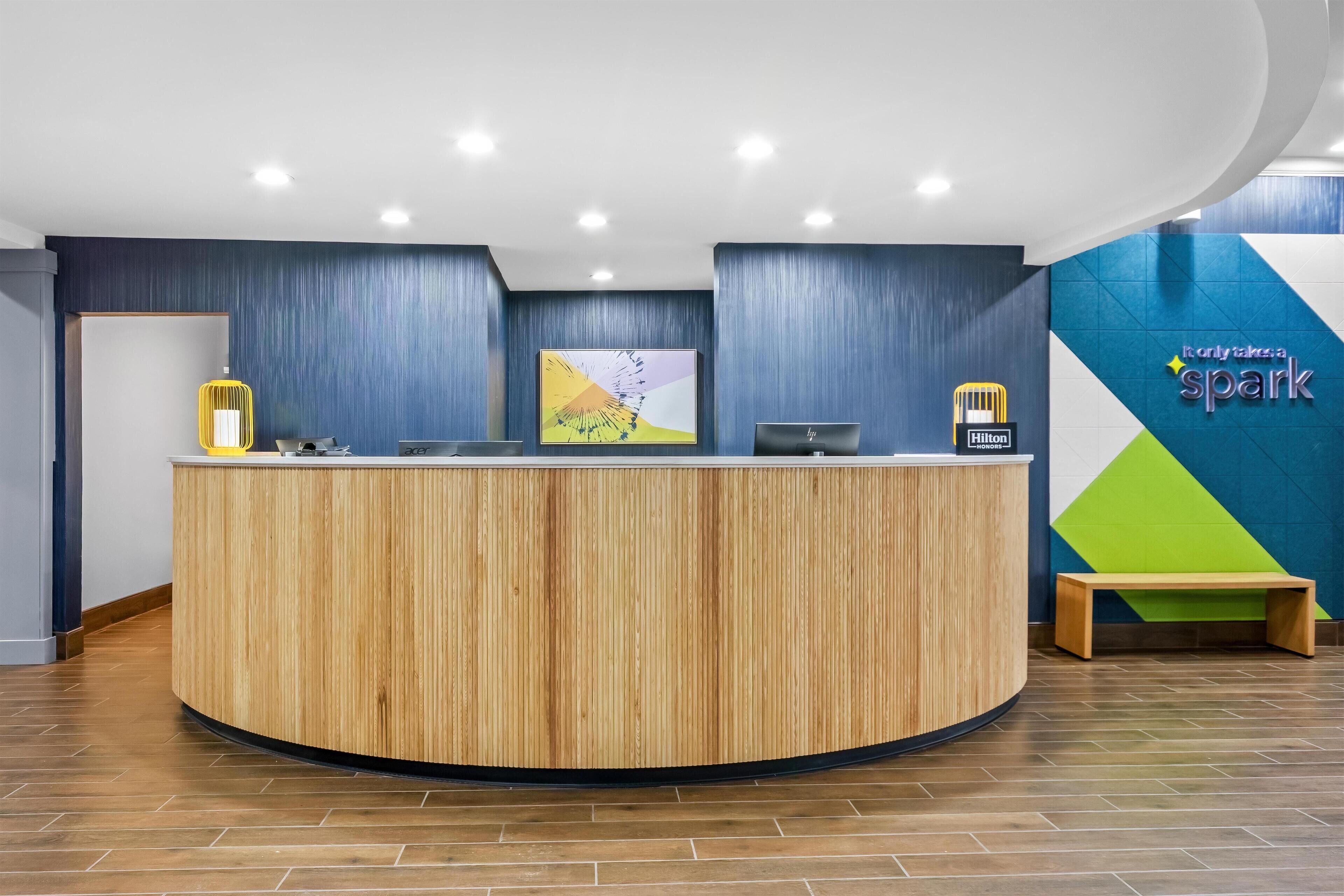 A reception desk with a wooden finish, a bench, and a wall with a geometric design and the text "It only takes a spark".