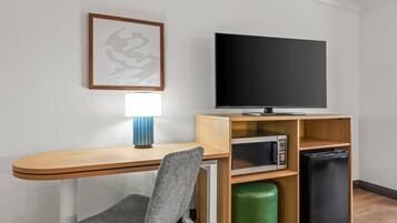 A hotel room with a wooden desk, a chair, a flat-screen TV, a microwave, and a refrigerator.