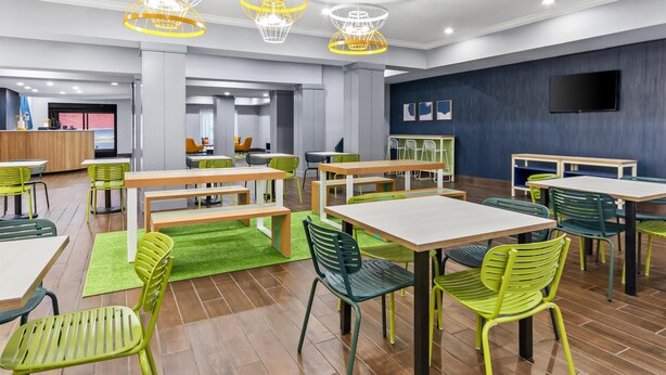 Een moderne eetruimte met groene en witte tafels en stoelen, een receptiebalie en een flatscreen televisie aan de muur.