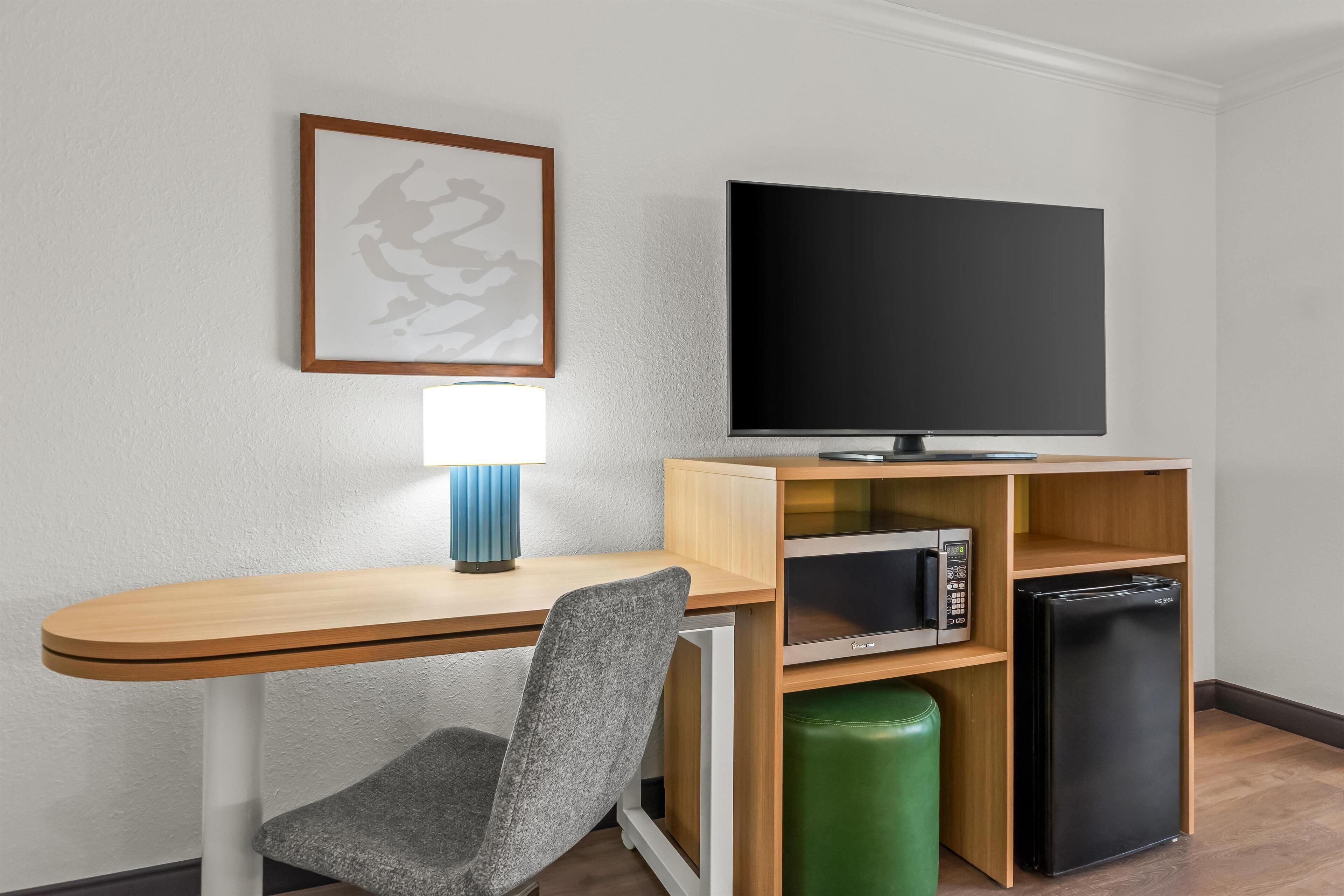 A hotel room with a wooden desk, a grey chair, a flat-screen TV, a microwave, a mini-fridge, and a trash bin.