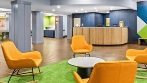 Een moderne hotel lobby met een receptiebalie, oranje stoelen, een ronde tafel en een groen tapijt.