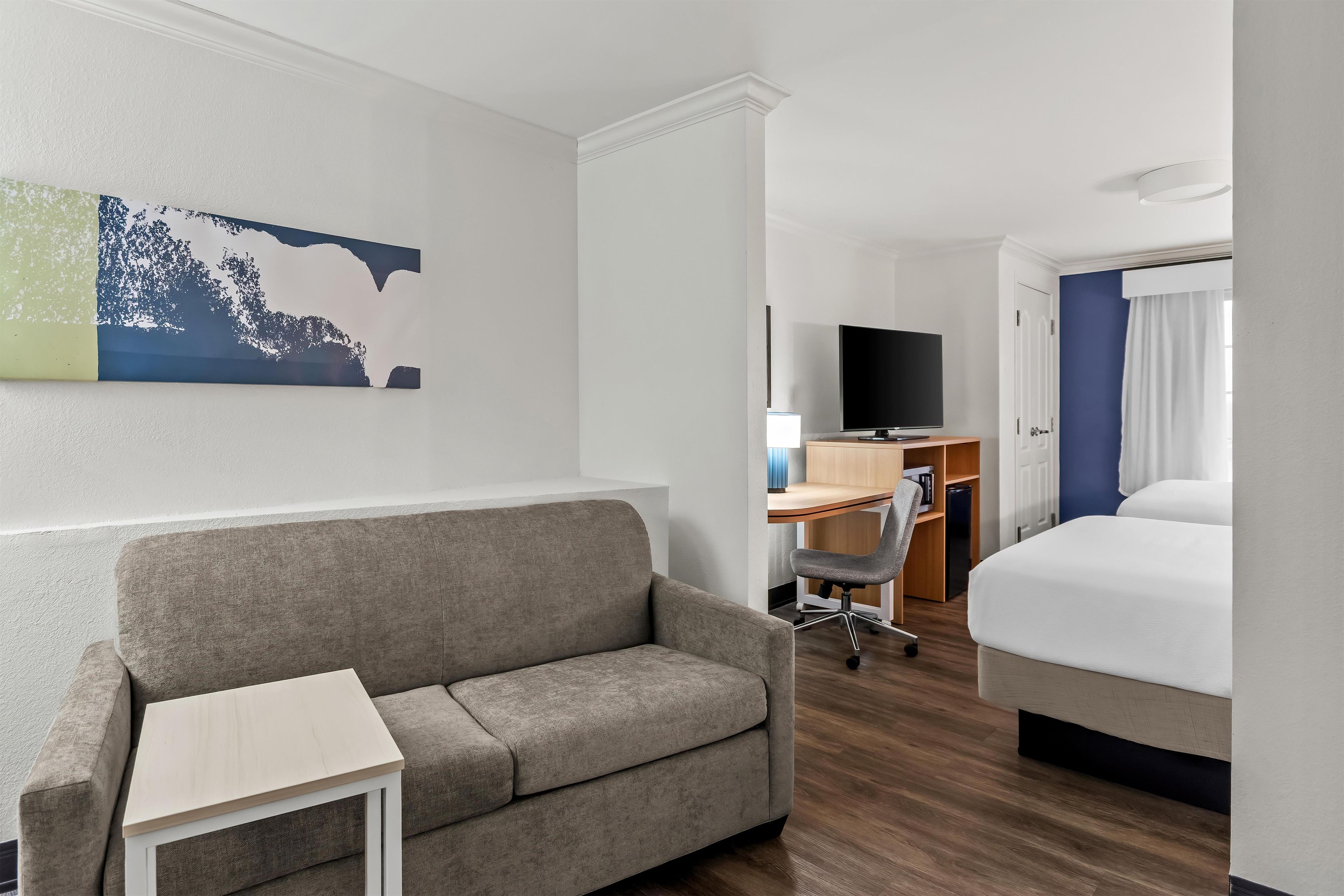 A hotel room with a sofa, a small table, a desk with a chair, a television, and a bed.
