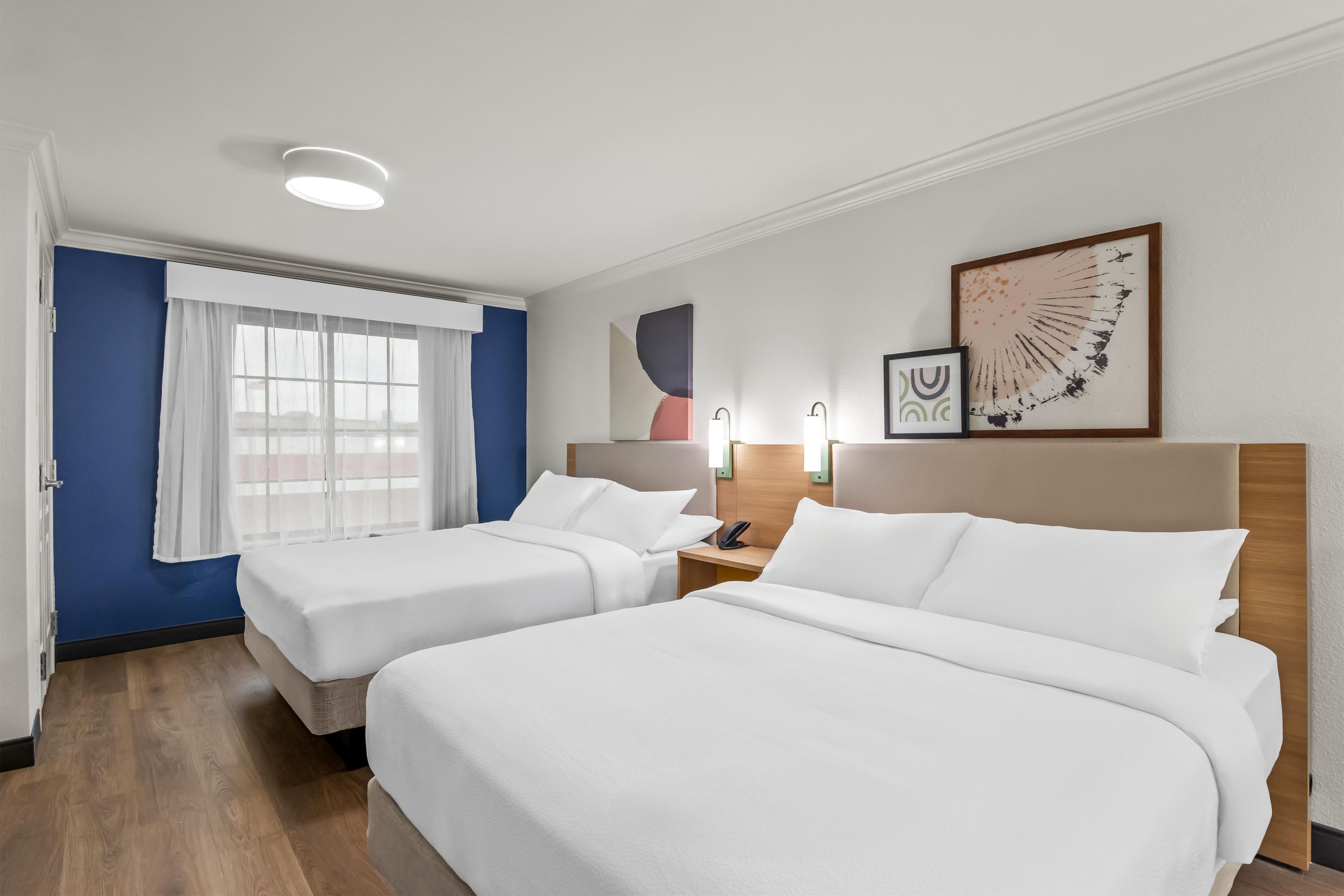 A hotel room with two beds, a window with sheer curtains, a wall-mounted artwork, and a bedside table with a phone.