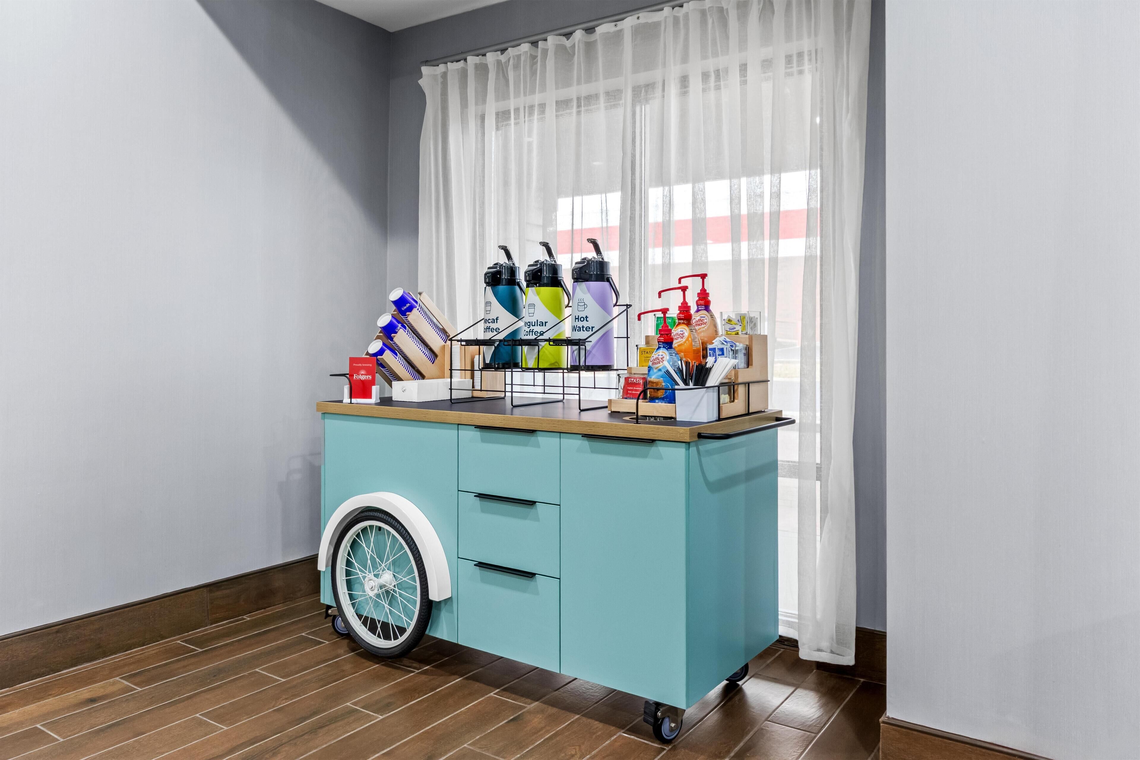 Un chariot mobile de couleur turquoise, doté d’une roue avant et rempli de divers articles, notamment des bouteilles et des récipients, est placé dans une pièce dont le sol est en bois et qui possède une fenêtre ornée de rideaux légers.