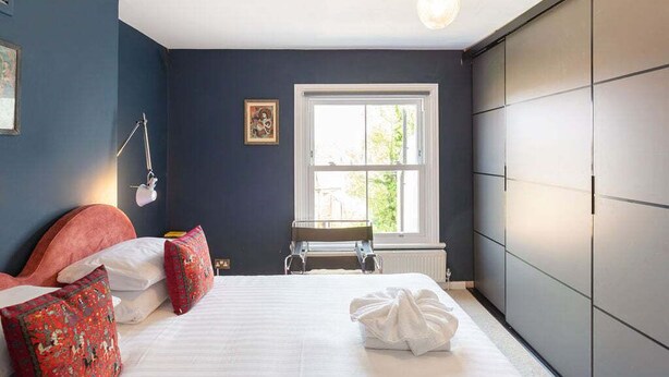 A bedroom with a bed, pillows, a bedside table, a lamp, a window, and a radiator.