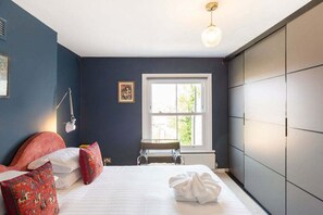 A bedroom with a bed, pillows, a bedside table, a lamp, a window, and a radiator. - GuestReady – 2bed near West Dulwich (London)