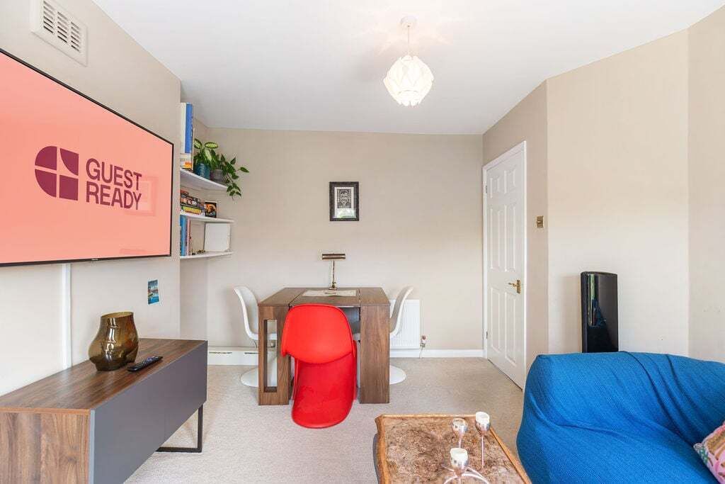 Een moderne woonkamer met een flatscreen televisie waarop “GUEST READY” staat, een houten salontafel, een blauwe bank, een rode fauteuil en een eettafel met witte stoelen.