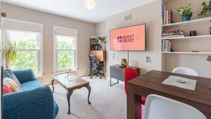 A modern living room with a large TV screen displaying "GUEST READY", a cozy blue sofa, a wooden coffee table, and a bookshelf filled with books and plants.