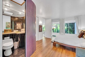 Ein modernes Schlafzimmer mit Holzboden, einem großen Bett, einem Fenster mit Vorhängen sowie einem Badezimmer mit Toilette und einer Fliesenwand.