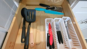 Un tiroir de cuisine contenant une planche à découper en bois, une spatule noire, un couteau bleu, un couteau à manche rouge et une râpe.