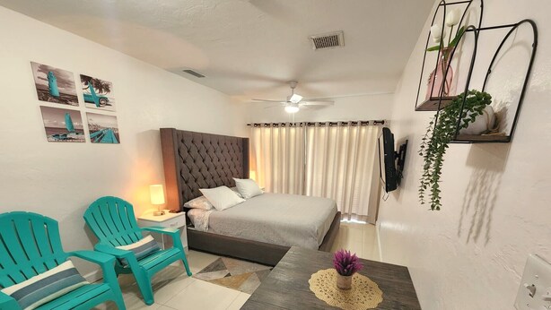 Une chambre d’hôtel avec un lit, deux fauteuils turquoise, une petite table ornée d’une fleur violette, un ventilateur de plafond, ainsi qu’un téléviseur et une plante fixés au mur.