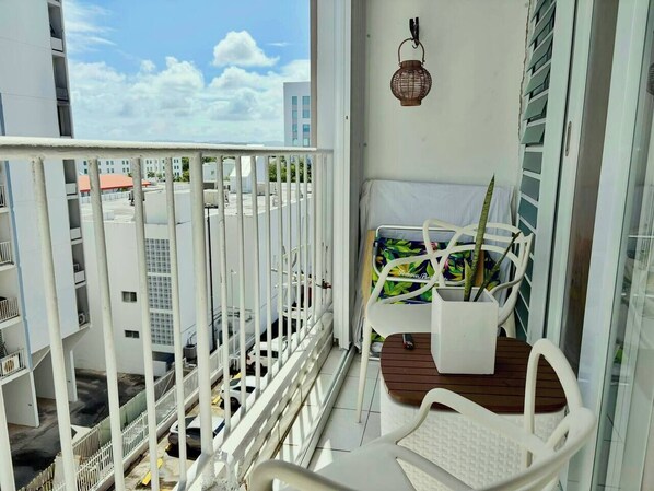 A balcony with a white railing, a white chair, a small table with a plant, and a view of buildings and sky. - Studio 3 minutes to the airport| Free Parking|Pool (Carolina)