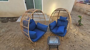 Two wicker swing chairs with blue cushions and a small table in a sandy outdoor area.