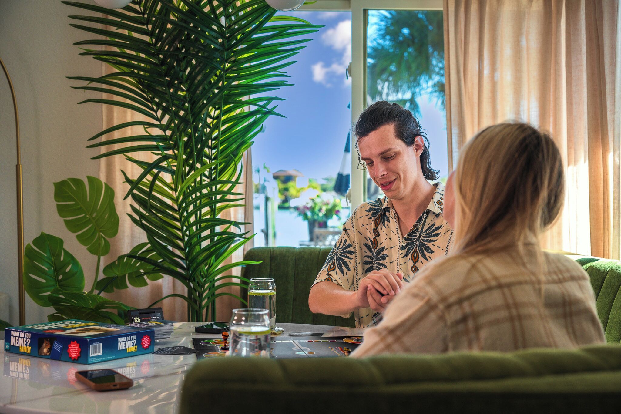 Une pièce avec une grande plante, une table sur laquelle est posé un jeu de société et une fenêtre avec des rideaux.