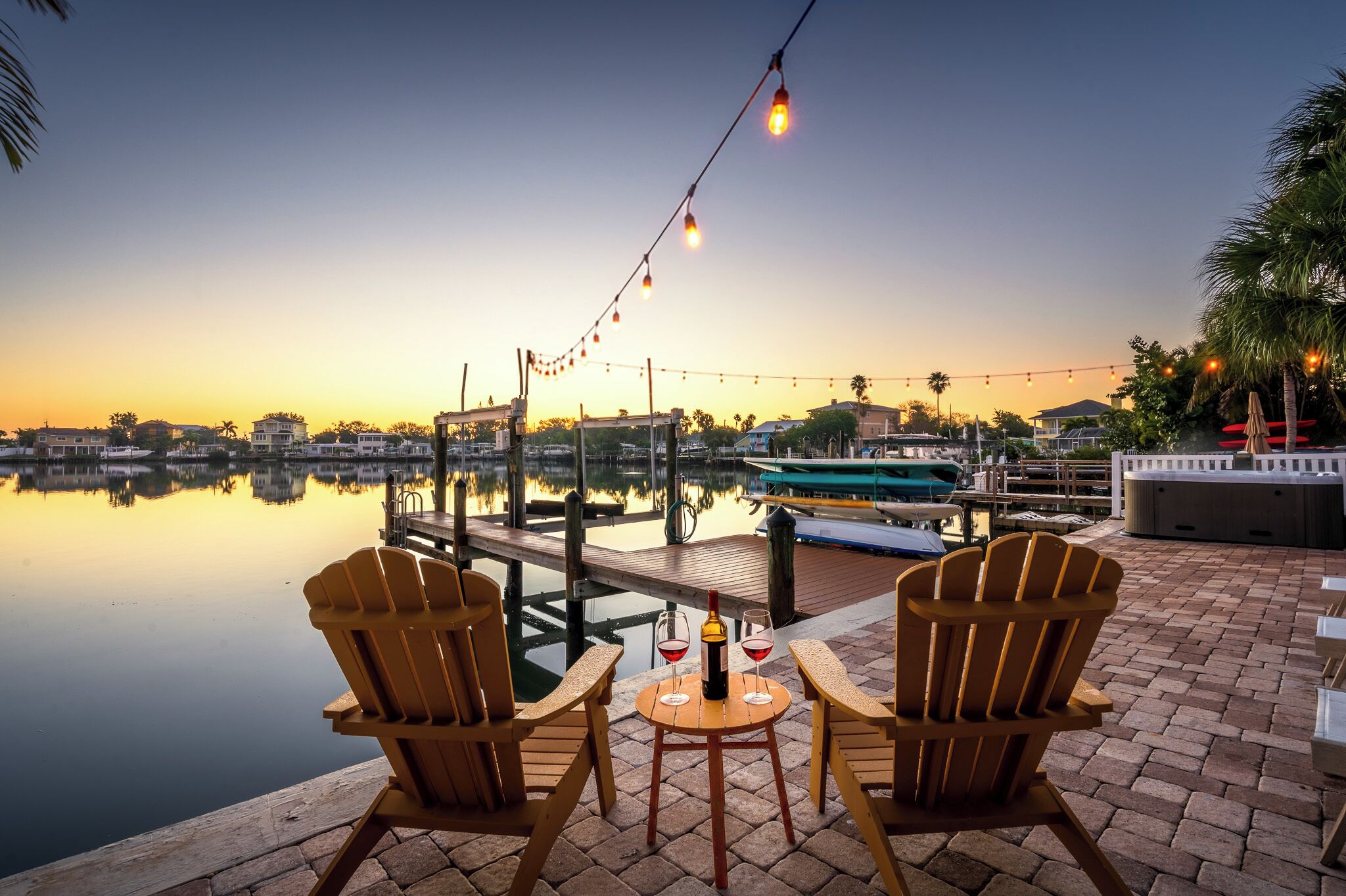 A waterfront patio with wooden chairs and a table, a dock, and a boat in the background.