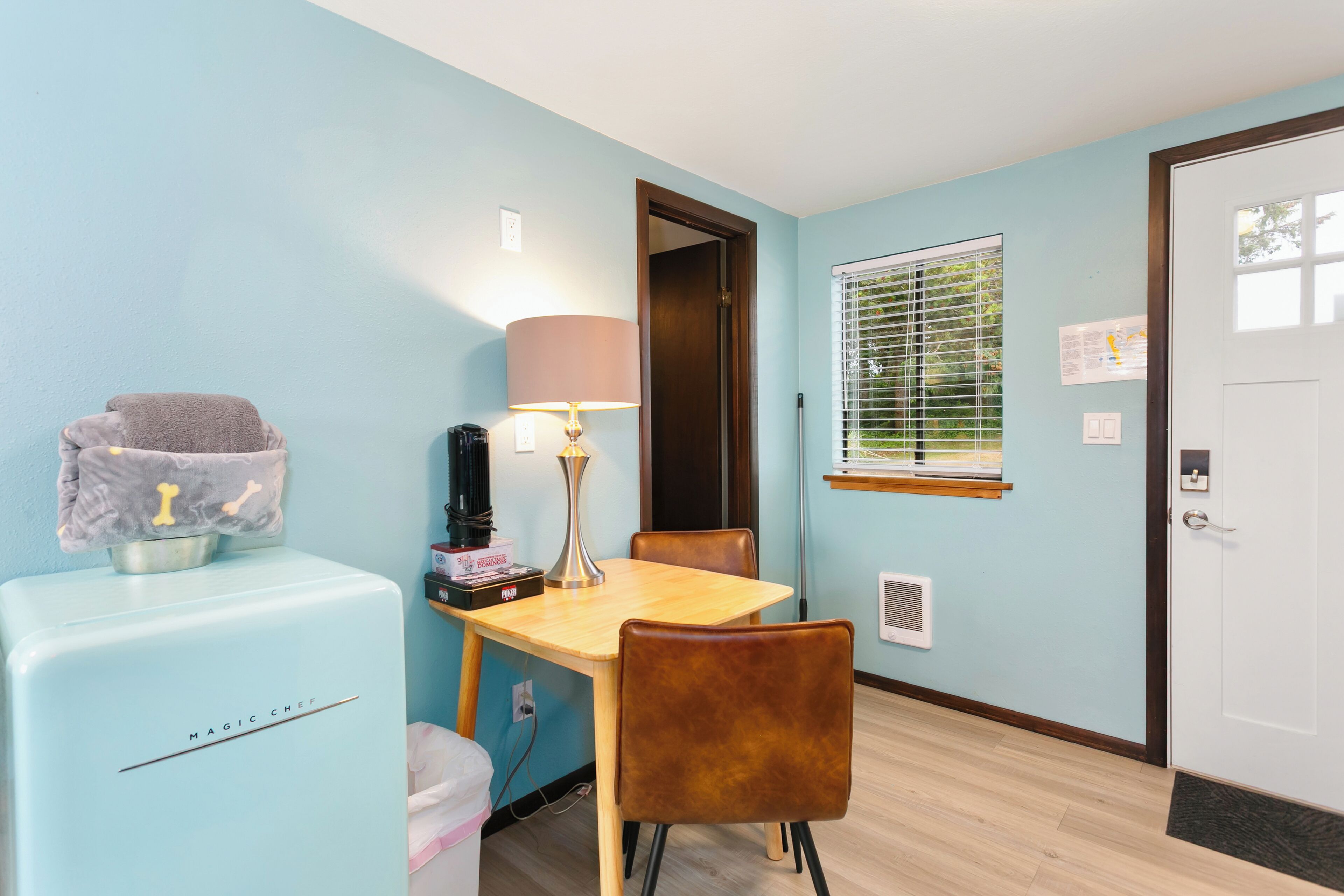 A compact room with a light blue wall, a wooden table, a brown chair, a small refrigerator, a lamp, and a window with blinds.