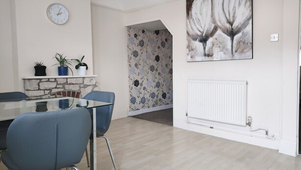 Een eetgedeelte met een glazen tafel, blauwe stoelen, een klok, potplanten, bloemenbehang en een radiator.