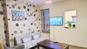 Een woonkamer met een bank, een salontafel, een aan de muur bevestigde televisie en een raam met planten op de vensterbank.