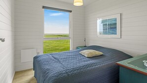 Une chambre avec une grande fenêtre, un lit recouvert d’une courtepointe, une table de chevet, une lampe et un tableau encadré au mur.