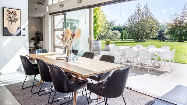 A modern dining area with a wooden table, black chairs, and a view of a garden through large glass doors.