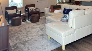 A spacious living room with a high ceiling, a white leather sofa, a brown armchair, and a dining area with a table and chairs.