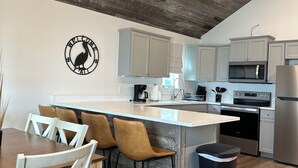 A modern kitchen with a wooden ceiling, white cabinets, a central island with bar stools, and a trash bin.