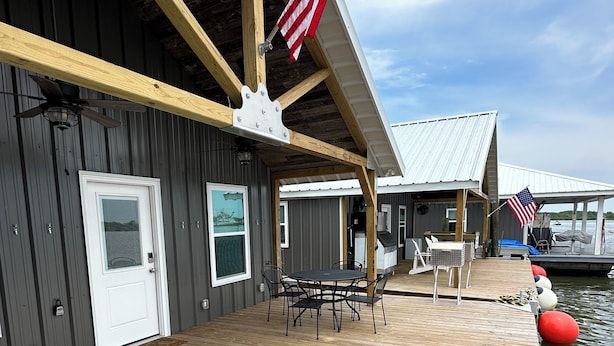 A wooden deck with a table and chairs, an American flag, and a dock extending into a body of water.